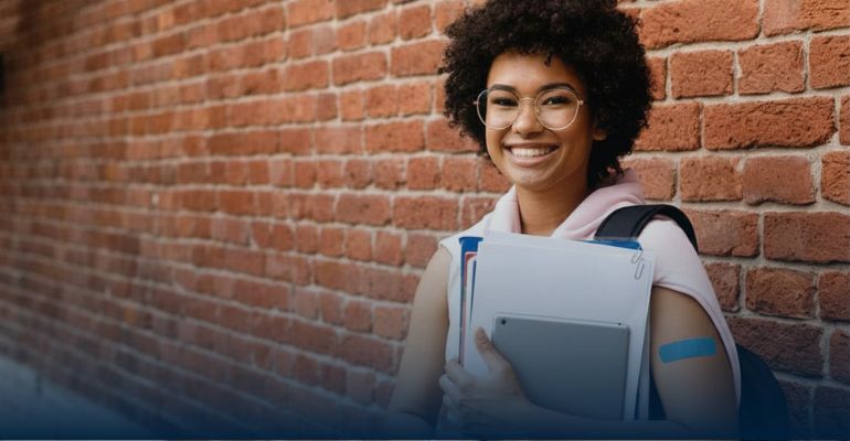 A young teen girl standing against a brick wall holding books smiling into the camera